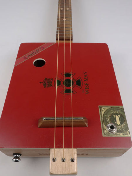 Front view of a Cigar Box Guitar handmade by Mike Snowden. This is a Red color cigar box guitar with The Wise Man logo printed on the front. 3 String Cigar Box Guitar with a sound hole and walnut bridge.