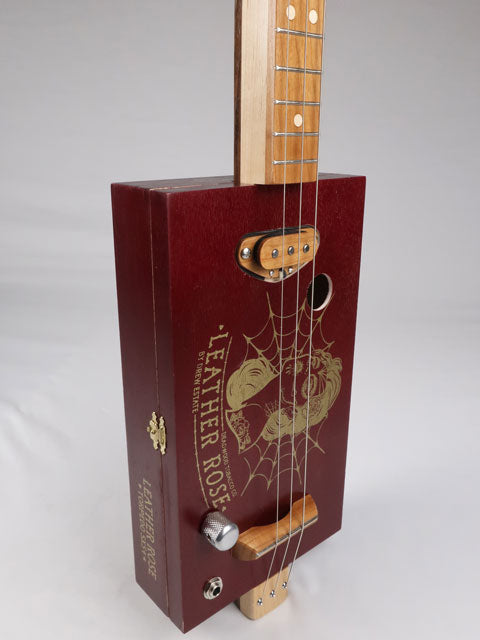 Tilted front view of a Leather Rose 3 string cigar box guitar. Red painted cigar box with gold Leather Rose graphic on the front. This cigar box guitar is handmade by Mike Snowden of Snowden Guitars.