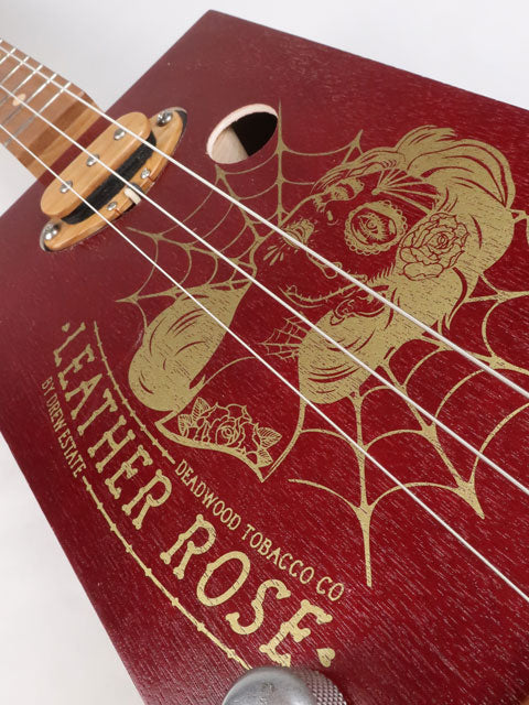 Close up tilted view of a Leather Rose 3 string cigar box guitar. Red painted cigar box with gold Leather Rose graphic on the front. This cigar box guitar is handmade by Mike Snowden of Snowden Guitars.