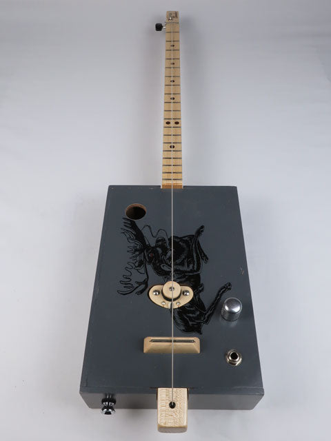 Front view of a one string diddly bow cigar box guitar with a moose on the front. This cigar box guitar is handmade by Mike Snowden and had a maple single coil pickup.