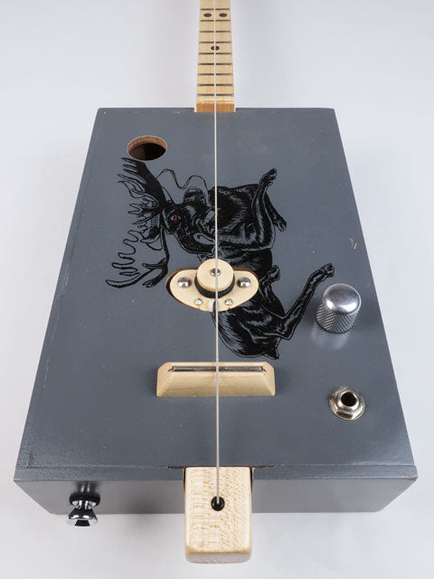 Front view of a one string diddly bow cigar box guitar with a moose on the front. This cigar box guitar is handmade by Mike Snowden and had a maple single coil pickup.