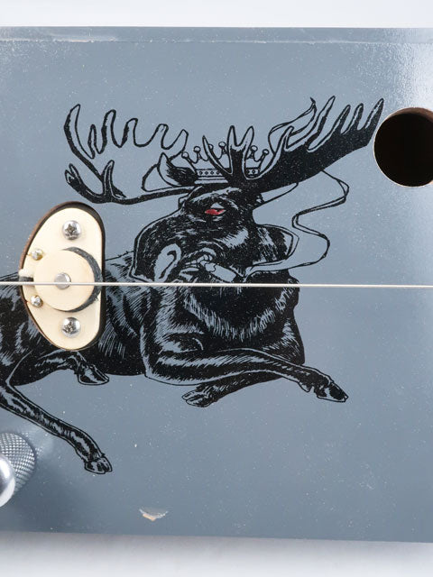 Close-up view of a one string diddly bow cigar box guitar with a moose smoking a cigar on the front. This cigar box guitar is handmade by Mike Snowden.